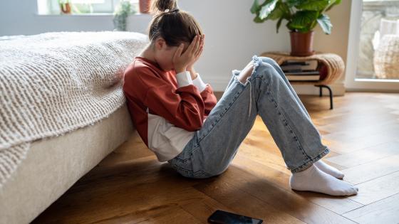 Teenage girl with head in hands sitting on floor next to mobile phone